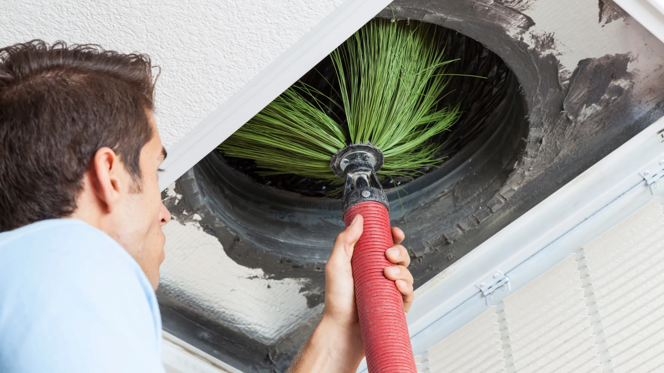 Person using a red vacuum hose with a brush attachment to clean an air duct, showcasing the importance of duct cleaning for improving indoor air quality.